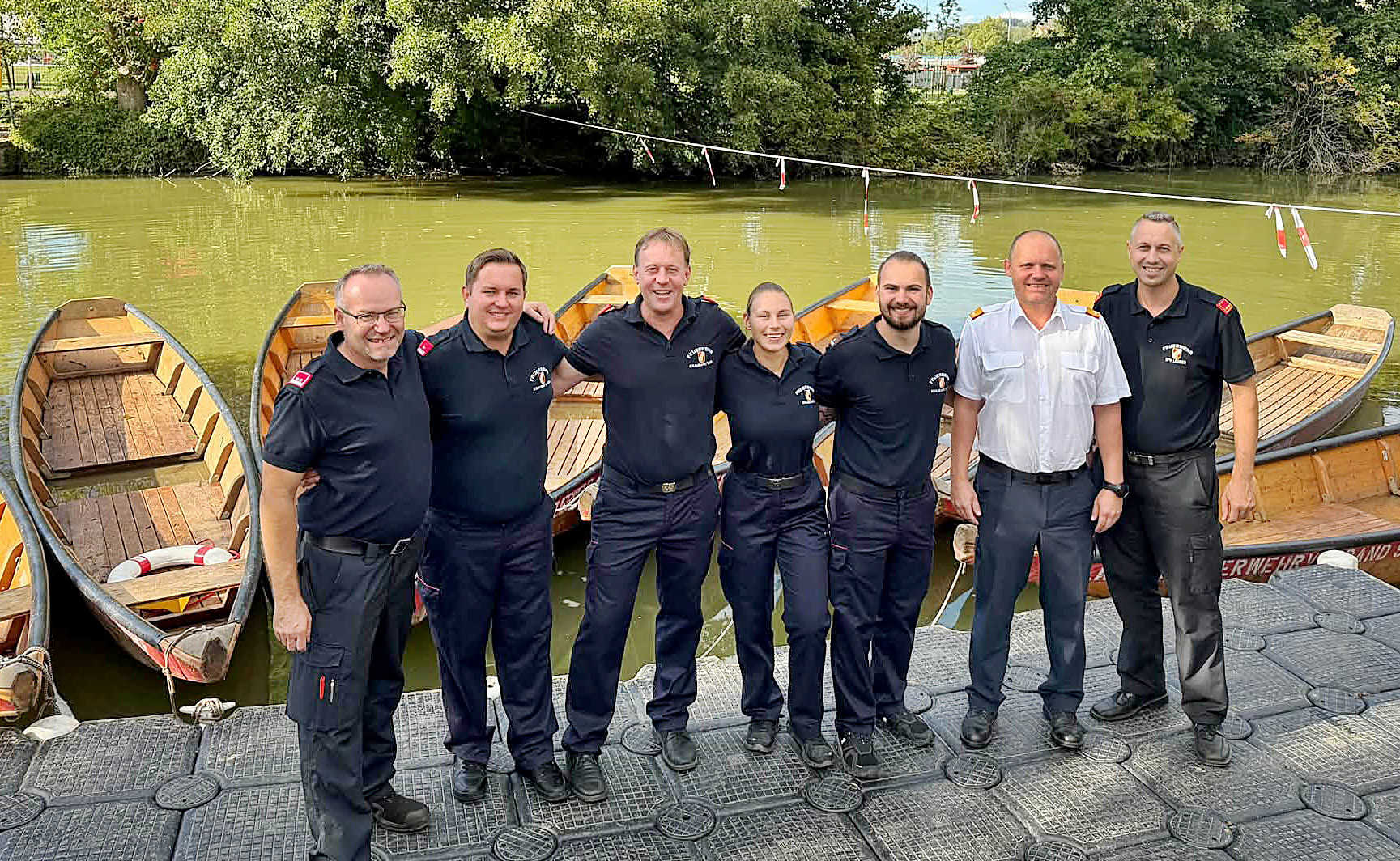 Feuerwehrleute in Uniform stehen am Ufer eines Flusses vor Ruderbooten
